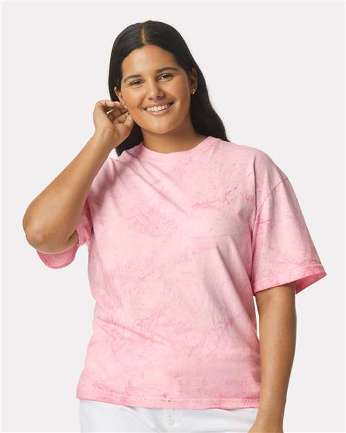 Person in a pink short-sleeve t-shirt with a marbled pattern, smiling with one hand raised near the ear.