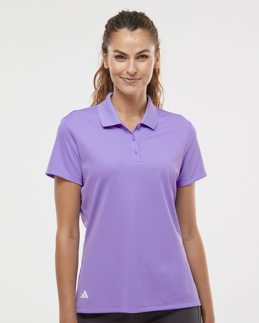 A woman modeling a short-sleeve purple polo shirt against a clean light gray studio background. The polo has a three-button placket and a small white Adidas logo near the lower left hem.