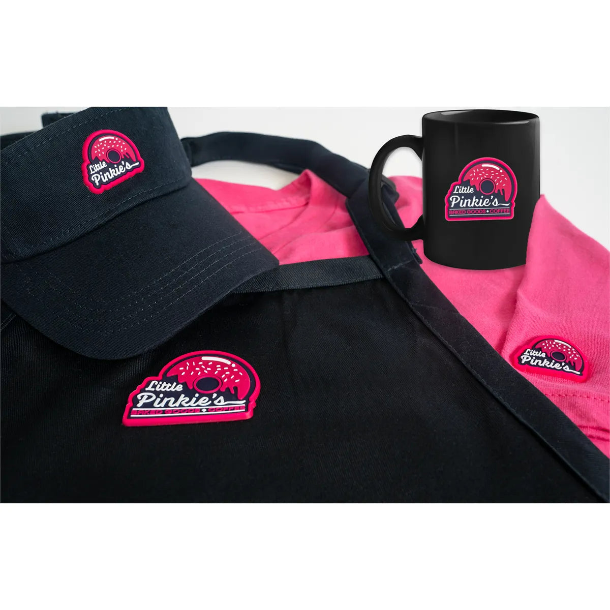Black cap, pink shirt, and black mug with 'Little Pinkie's' logo on a white background