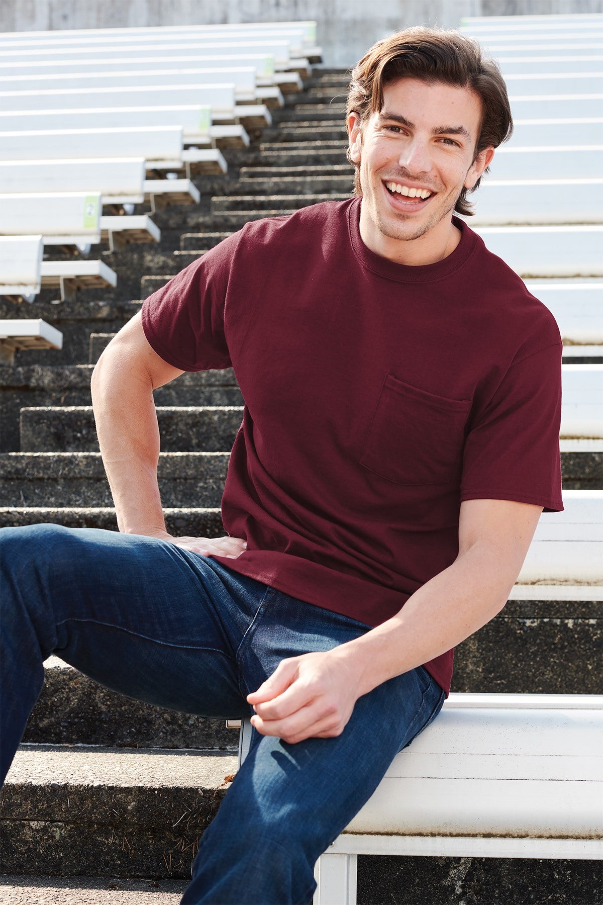Man sitting on a bench wearing a maroon t-shirt and blue jeans, smiling.